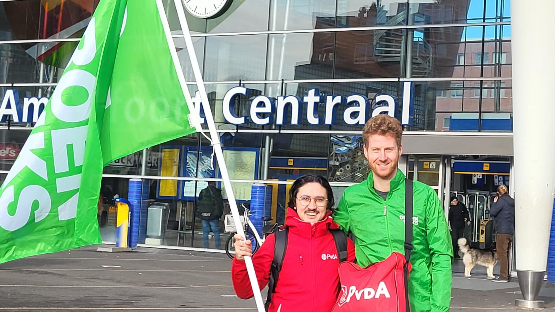 Twee jongemannen, de een met rood jasje en groene vlag, de ander met groen jasje. Arm in arm.